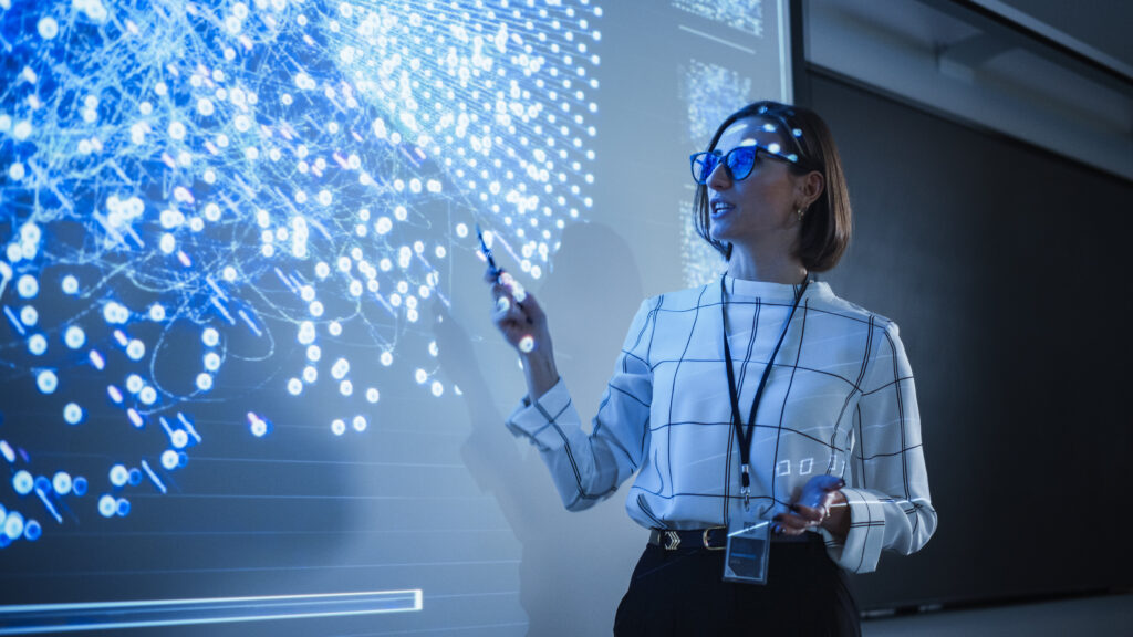 A female teacher teaches computer coding in a darkened classroom.