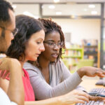 Three educators look at the same computer screen in a computer lab.